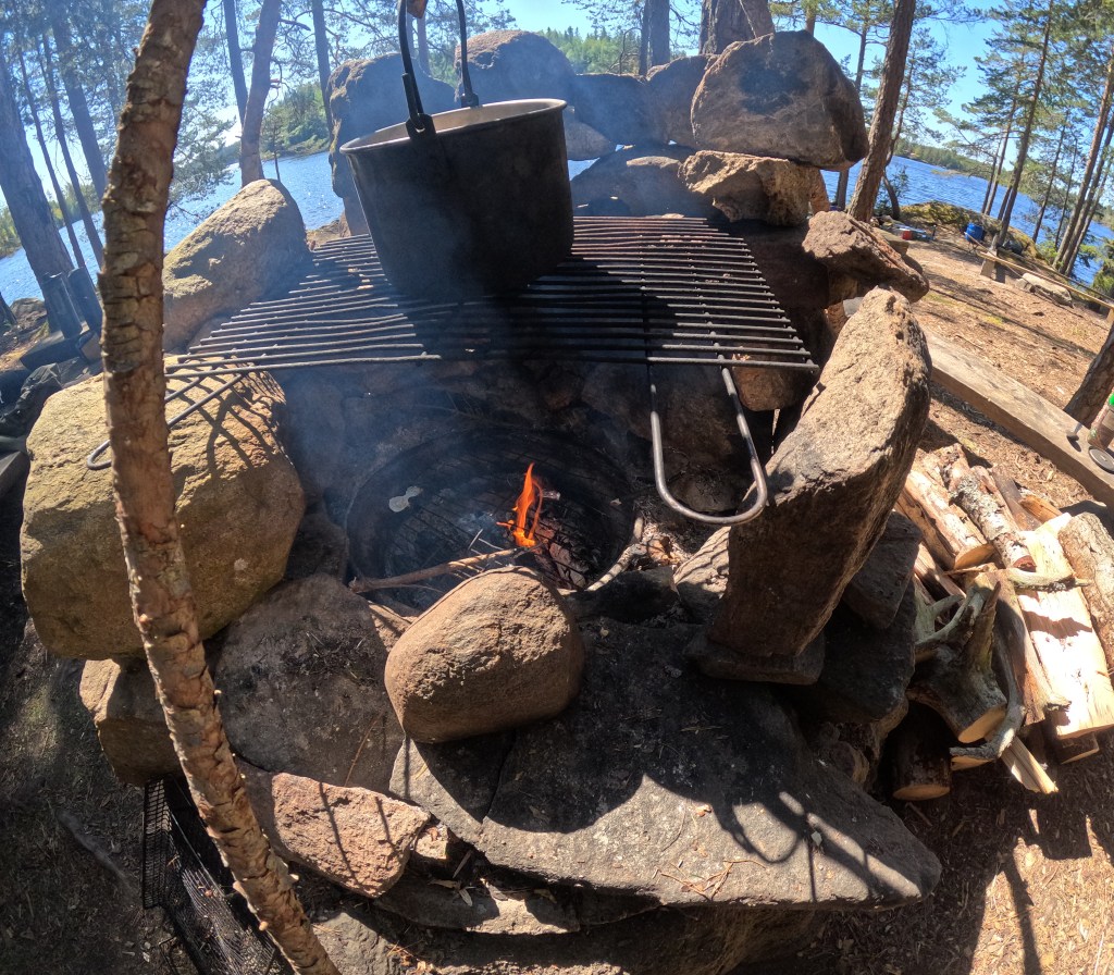 Ein Lagerfeuerplatz in der Natur mit einem Gitterrost und einem Topf über dem offenen Feuer, umgeben von Steinen und Bäumen, mit einem See im Hintergrund.